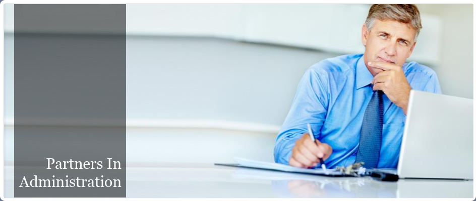Partners in Administration. Image of a man at a desk in front of a laptop, writing on a clipboard with a pen and looking at the camera with his other hand on his chin as if he is listening to you.
