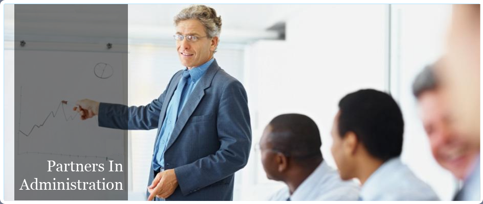 Partners in Administration. Image of a man giving a presentation in front of a chart. He is pointing at the chart and there are four other men sitting at a long desk looking at his presentation.