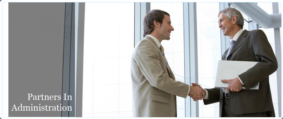 Partners in Administration. Image of two men in business suits smiling and shaking hands with each other in front of white windows inside of an office building. The man on the right is holding a legal pad.